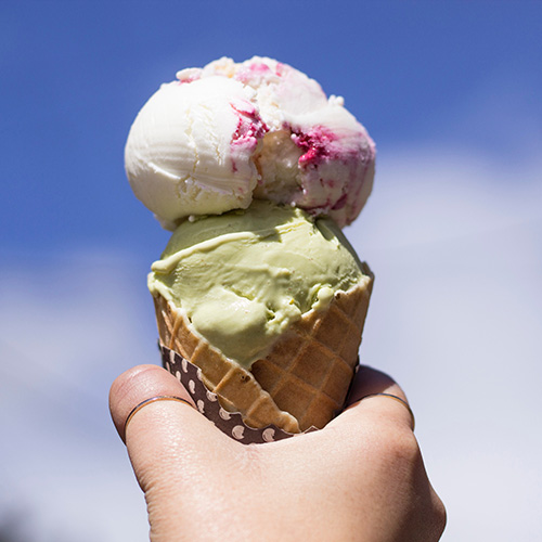 A hand holds up an ice cream cone against a blue sky with puffy white clouds.