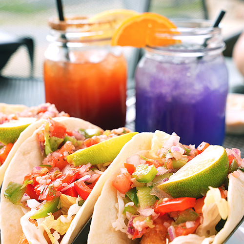 Two tacos sit on a table with cocktails.