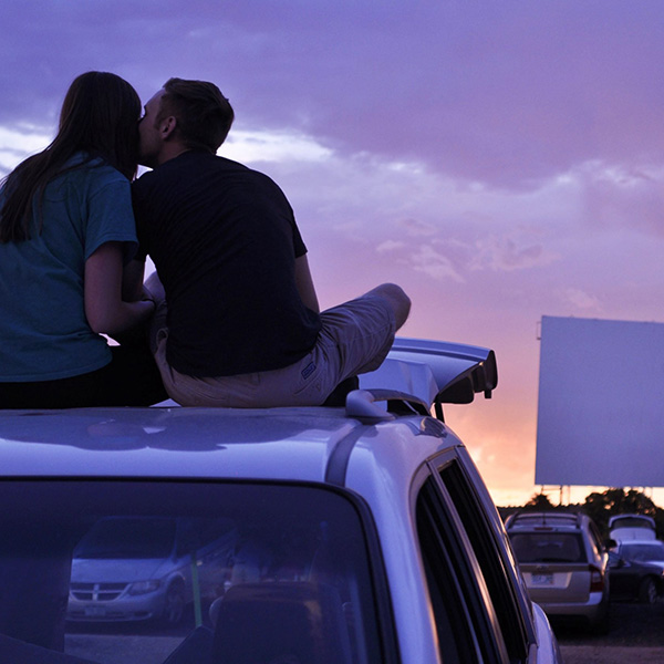 Two people sit on top of a car at the drive-in movies.